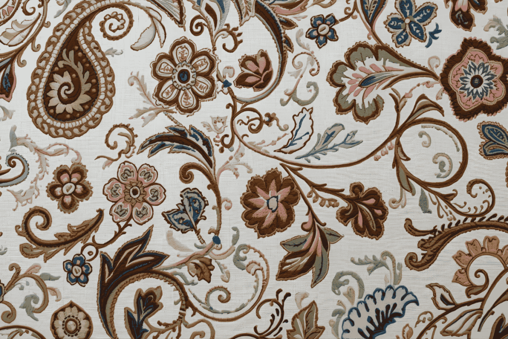 An intricately embroidered white fabric featuring a colorful paisley and floral pattern with swirling lines and leaves in shades of brown, blue, pink, and green.
